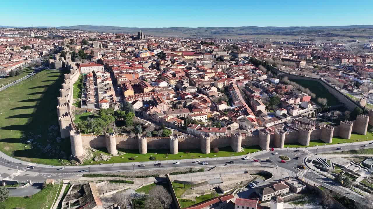 voo com uma câmera girando visualizando com um céu azul a cidade de ávila, um local de patrimônio mundial da unesco com sua parede de pedra e suas torres e uma estrada perimetral com veículos circulando na espanha