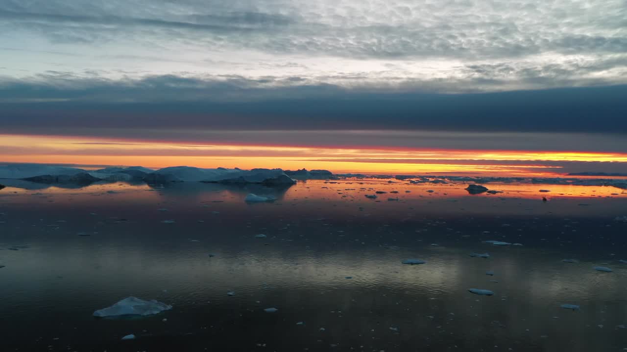 Aerial drone view of a stunning sunrise over frozen sea ice and calm Arctic waters in Greenland, showing warm orange light reflecting on icy surface beneath dramatic clouds