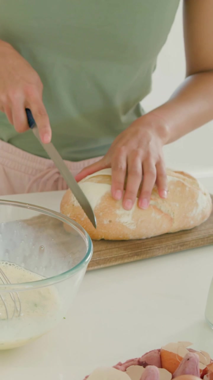 Vertical video: Arm reaching, woman in green whisking batter slicing loaf at island preparing meal