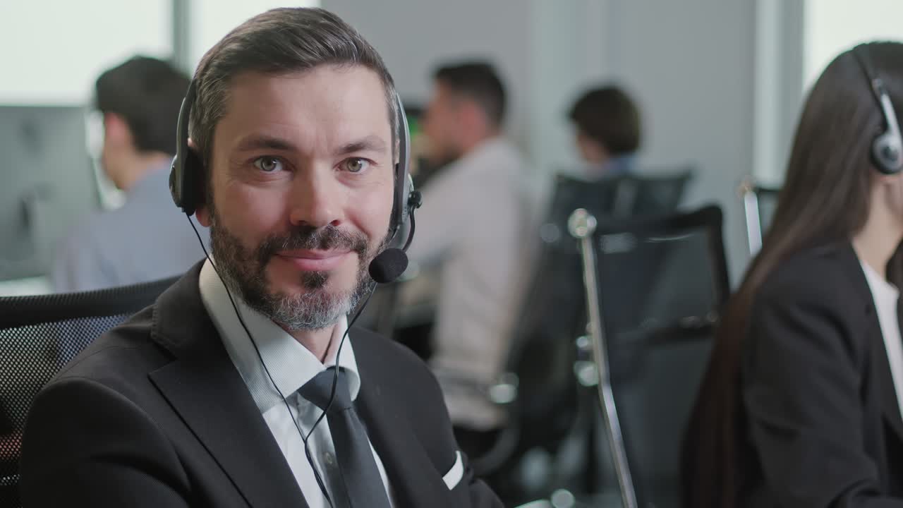 Portræt af en glad og smilende mand, der kigger ind i kameraet, mens han arbejder i et callcenter. Han bruger en stationær pc ved et computerbord og en telefonlinje i et moderne co-working-miljø.
