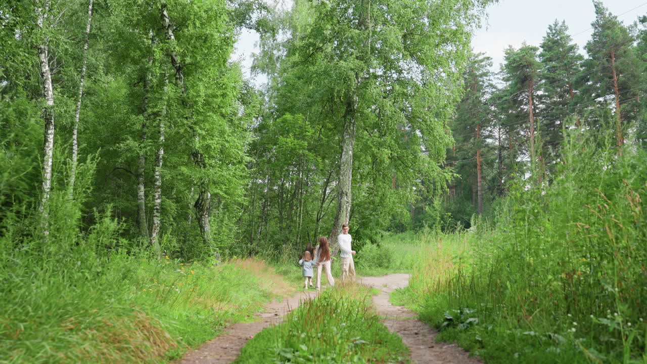 Long shot of little girls joyfully running after big brother along forest path surrounded by tall green trees and summer foliage, capturing playful sibling moment in bright natural light