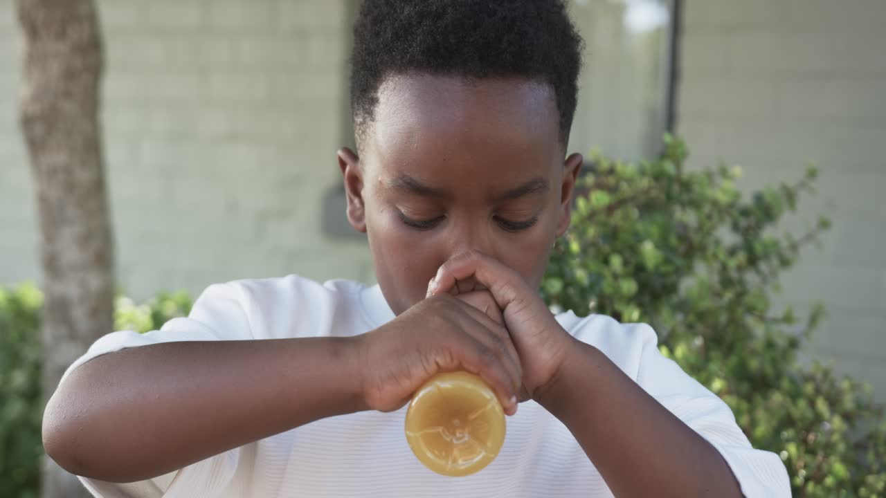 Joven africano bebiendo de una botella con boquilla naranja, luego sujetándola e inclinándola para terminar de hidratarse en el patio.