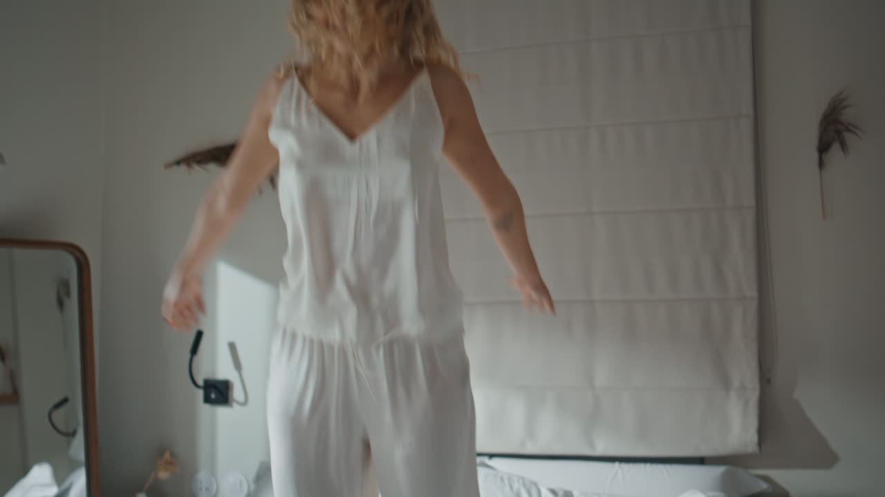 Happy woman dancing bed in morning. Joyful curly lady falling down in pajamas