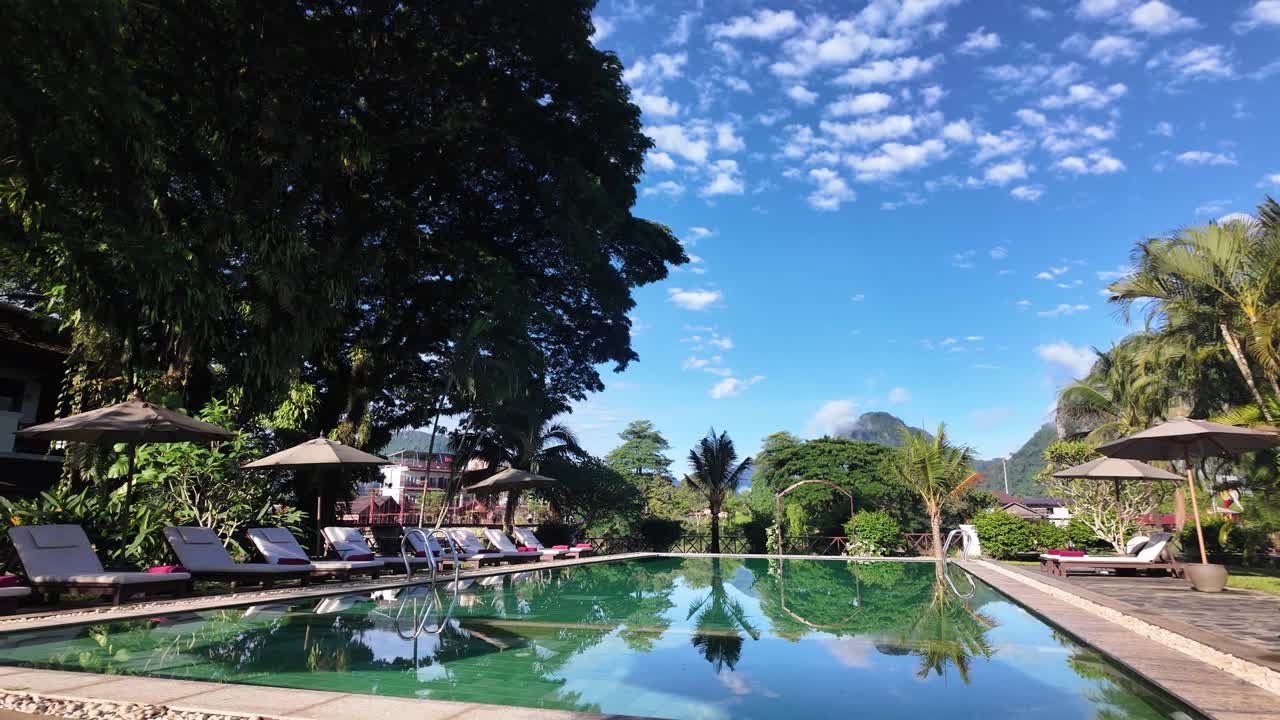 View of a swimming pool at a boutique resort in Vang Vieng Laos featuring loungers umbrellas palm trees and mountain views under a blue sky with clouds