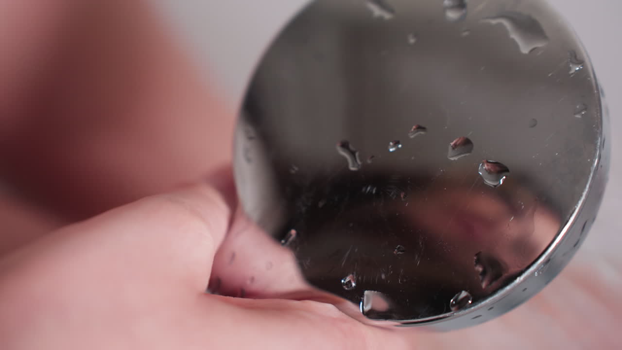 Close macro view hand holding handheld shower head with water droplets, mirrorlike chrome surface reflecting blurry face, soft focus background, hygiene scene in bathtub