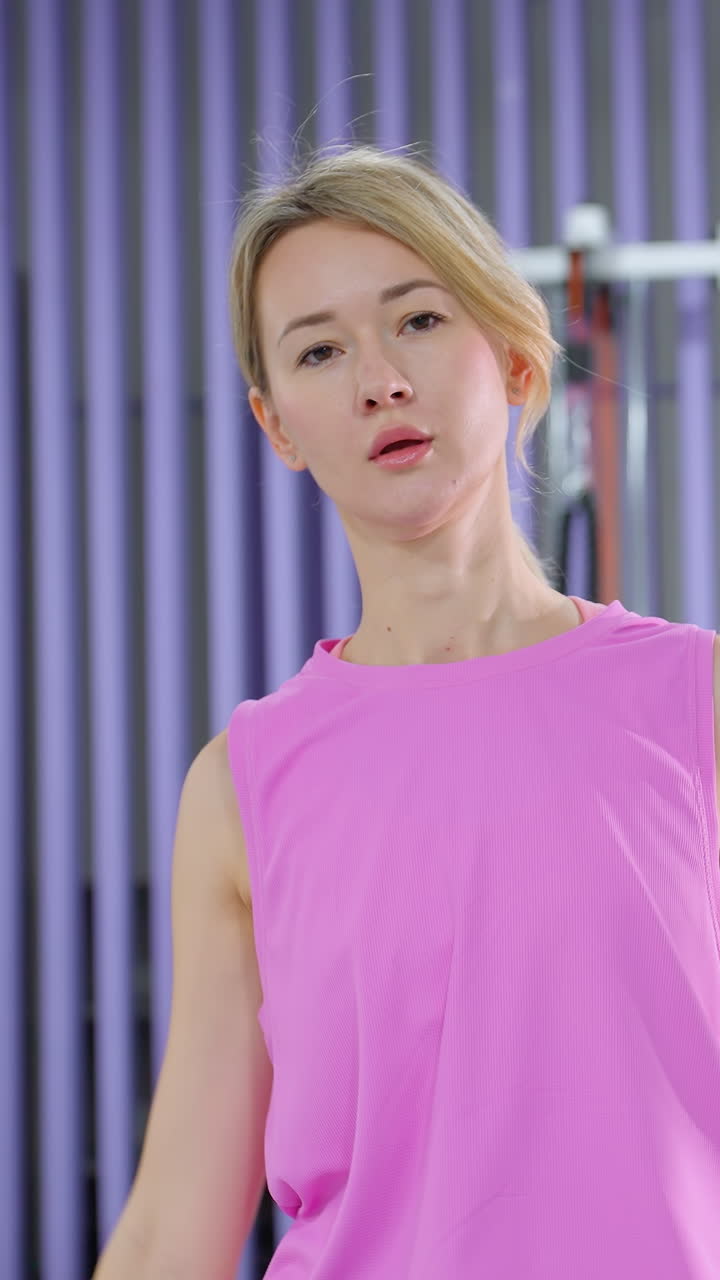 분홍색 스포츠 의류를 입은 여자가 체육관에서 오른쪽에서 왼쪽으로 팔을 펴고, 유연성과 형태를 보여 주며, 배경에는 다른 개인의 부분적인 시각이 있습니다.