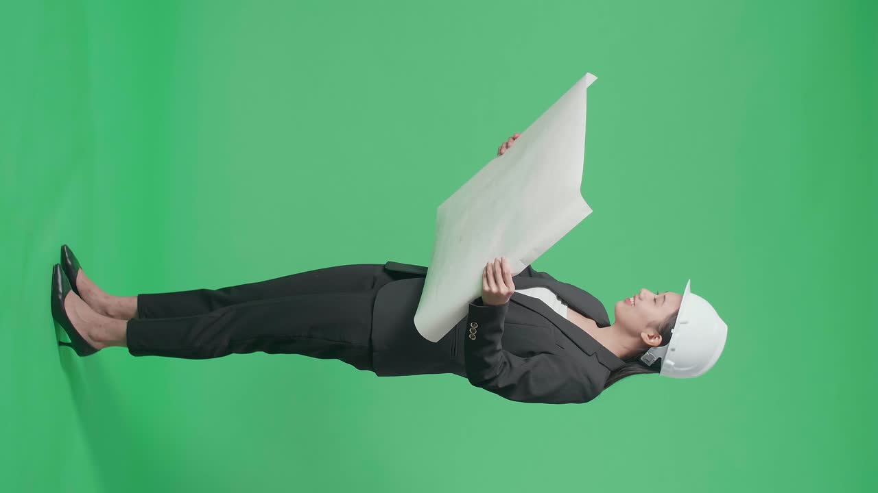 Full Body Side View Of Smiling Asian Female Engineer Looking At Blueprint Before Looking Around And Shaking Her Head In The Green Screen Studio