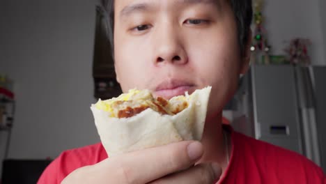 portrait of young man eating showing delicious scrambled egg and sausage burrito