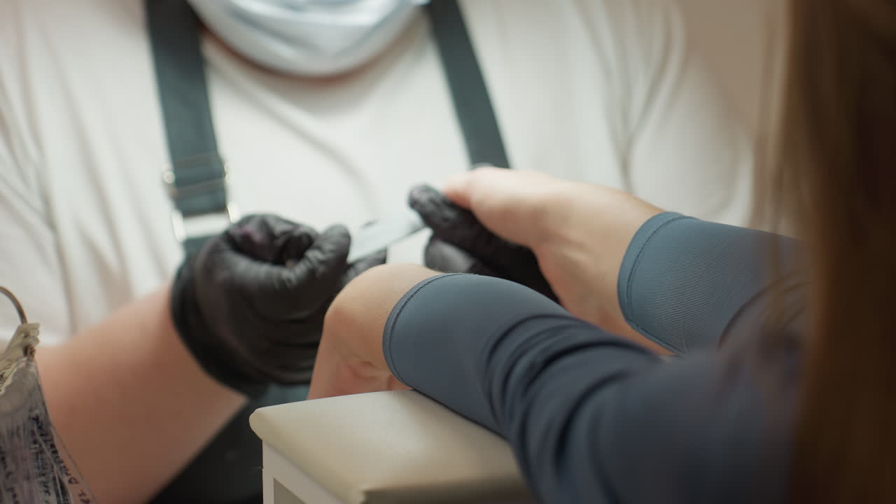 Nail technician wearing black gloves and protective mask shaping customer s nails using hand filer during manicure session in salon, focused view on interaction and detail-oriented nail care process