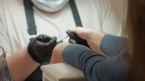 nail technician wearing black gloves and protective mask shaping customer s nails using hand filer during manicure session in salon, focused view on interaction and detail-oriented nail care process