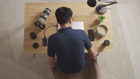 top view of asian male editor walking into the workspace sitting down and using a laptop next to the camera editing the video at home