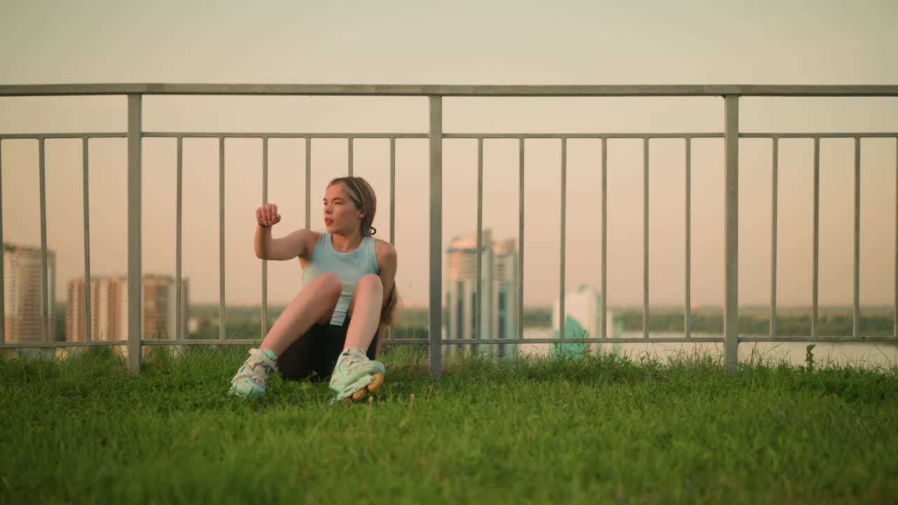 joven en patines sentada en un terreno cubierto de hierba, descansando con la mano en la pierna, disfrutando de la puesta de sol nocturna con el paisaje urbano borroso en el fondo