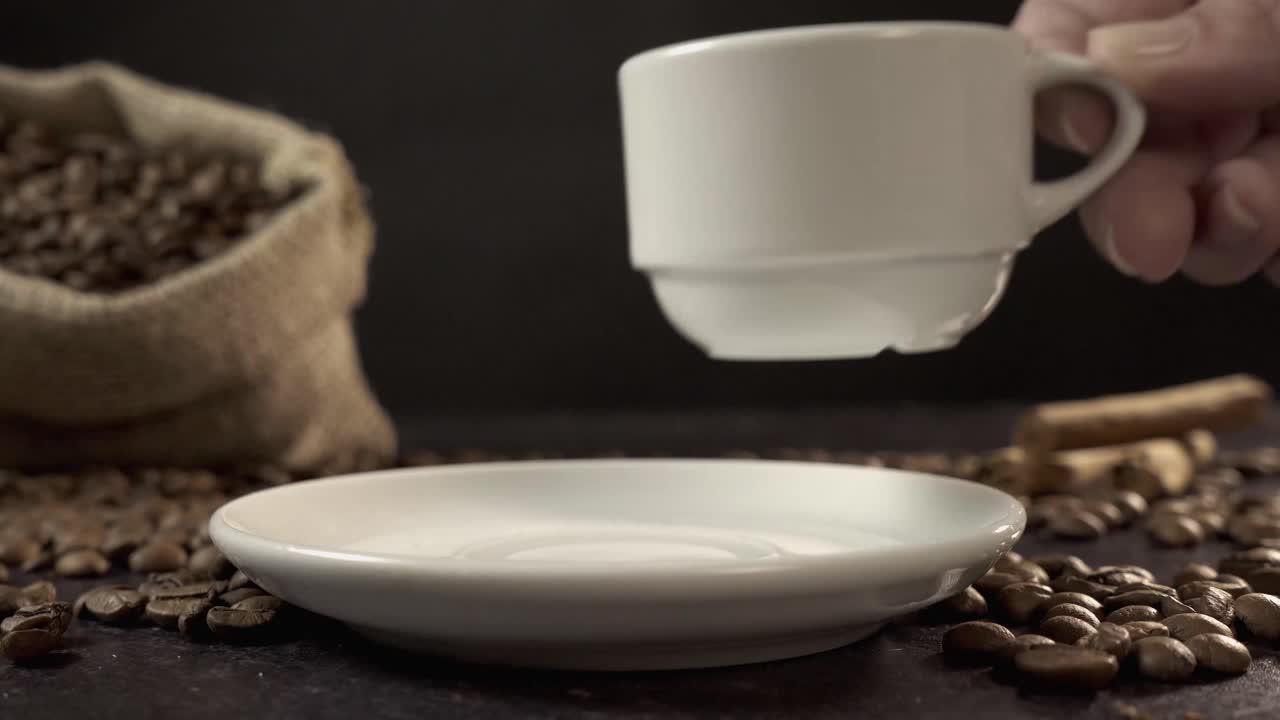 Hænder placerer hvid kaffekop på underkop midt i kaffebønner