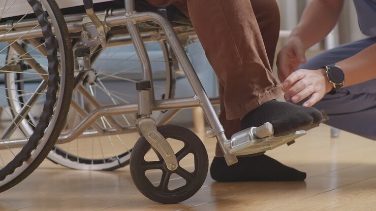 Close Up Of Male Nurse'S Hands Helping A Patient In Wheelchair Putting Feet On Footrest And Pushing Him To Another Room After A Physical Therapy At Home