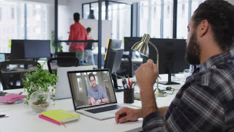 hombre de negocios de raza mixta sentado en un escritorio usando una computadora portátil haciendo una videollamada con un colega masculino