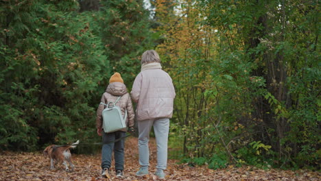 back view of child holding dog leash walking beside mother along forest path filled with autumn leaves, surrounded by vibrant green and orange foliage during peaceful nature stroll