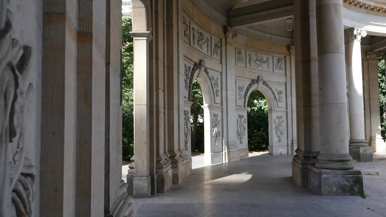 Sunlight falls across stone arches of Kolonnadenhof Colonnade on Museum Island as carved pillars and detailed walls create calm historic scene surrounded by greenery in central Berlin