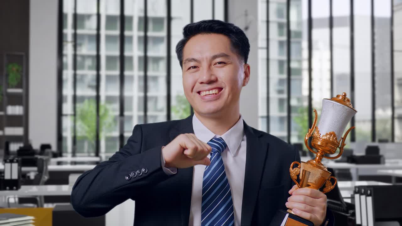 Close Up Of Asian Business Man In A Suit And Tie With A Gold Medal Showing Gold Trophy To Camera, Smiling, And Pointing Himself Being Proud Being Winner In Office