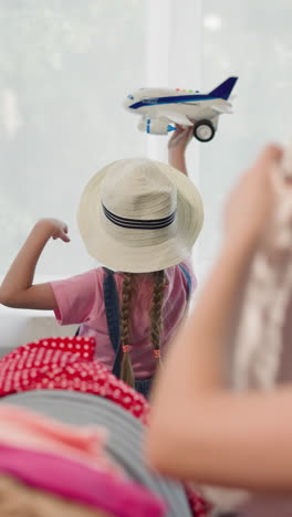 little daughter with straw hat plays with airplane while woman folds belongings on sofa in apartment. family journey preparation at home