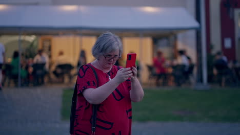 Older-woman-with-grey-hair-wearing-glasses-and-a-red-T-shirt-taking-pictures-with-her-smartphone-in-the-historical-city-Bardejov-in-Slovakia-during-a-blue-hour-at-dusk-in-slow-motion