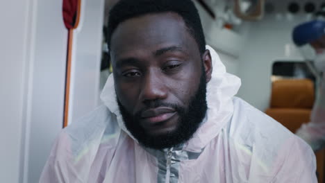 black paramedic in protective clothes looking at camera bearded african american doctor in hazmat suit blinking sleepily looking at camera while riding 911 service vehicle during pandemic