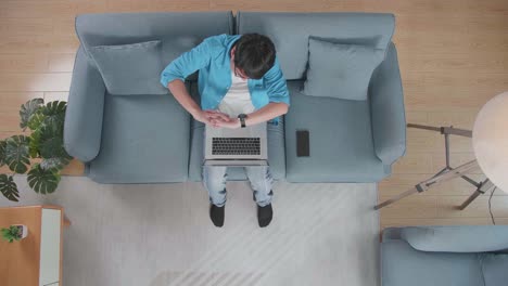 top view zoom out shot of a man stretching while sitting on the sofa with a smartphone typing on a laptop at home