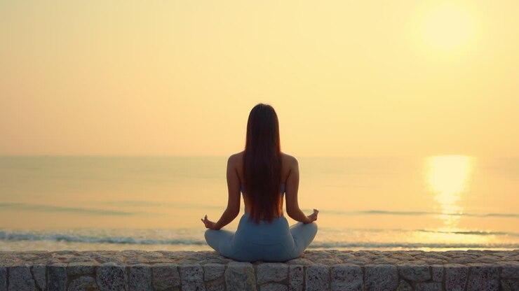 Mujer esbelta practicando Yoga, meditando en posición Zen cerca del océano durante una puesta de sol dorada.