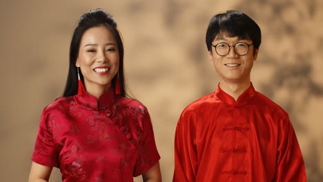 happy young asian couple in red traditional clothes looking at each other and smiling cheerfully at camera