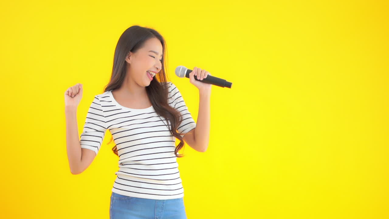 Beautiful young Asian woman singing with the microphone on yellow studio background