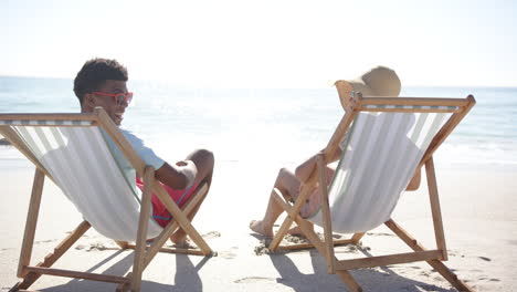 un joven biracial y una joven caucásica se relajan en sillas de playa junto al mar.