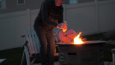 older man joins his wife by a fire pit on a cold night
