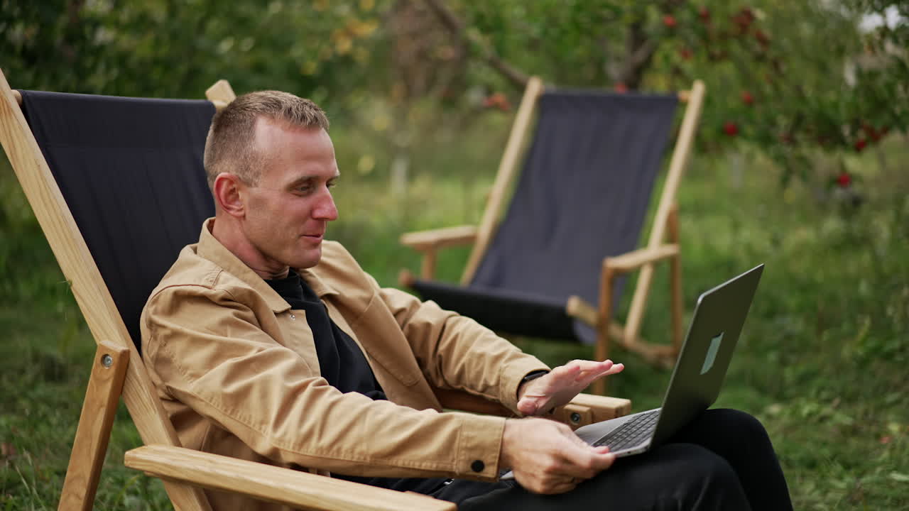 Emotional talk through the video chat on laptop. Man holding computer on his laps sitting in a garden chair outdoors. Apple garden blurred backdrop.