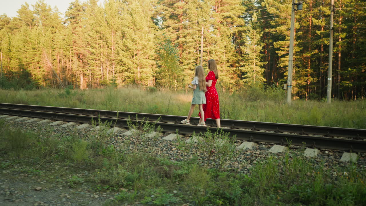 young adult and child walking side by side on rural railroad track during golden hour, surrounded by tall grass and forest trees, peaceful evening scene with power poles visible