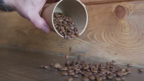 Mano-Derramando-Granos-De-Café-En-Una-Taza-Sobre-Una-Mesa-De-Madera