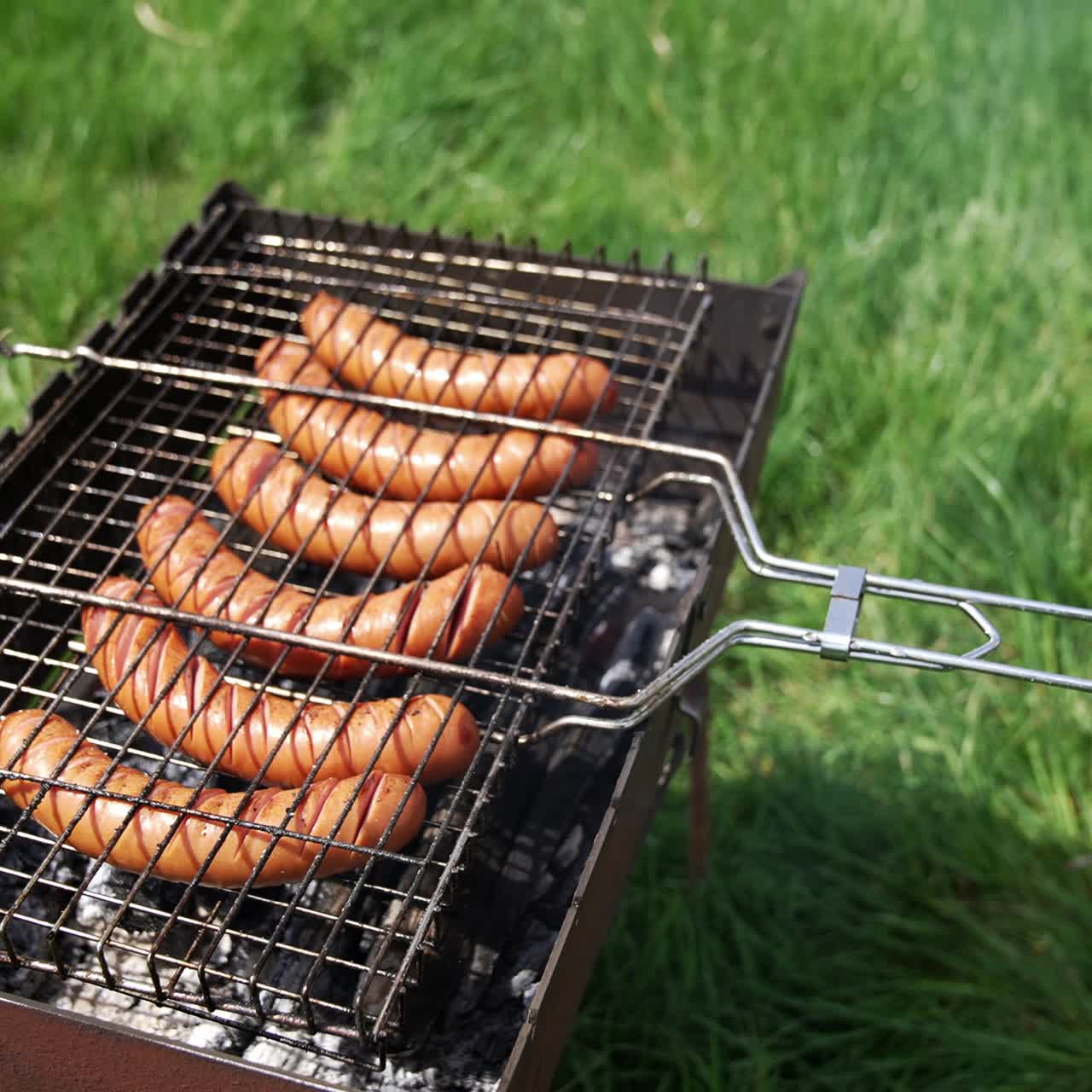 Tasty juicy sausages grilling over a fire in smoke. Hot dog sausages on a grill grate cooking on the green grass background. Barbeque in summer