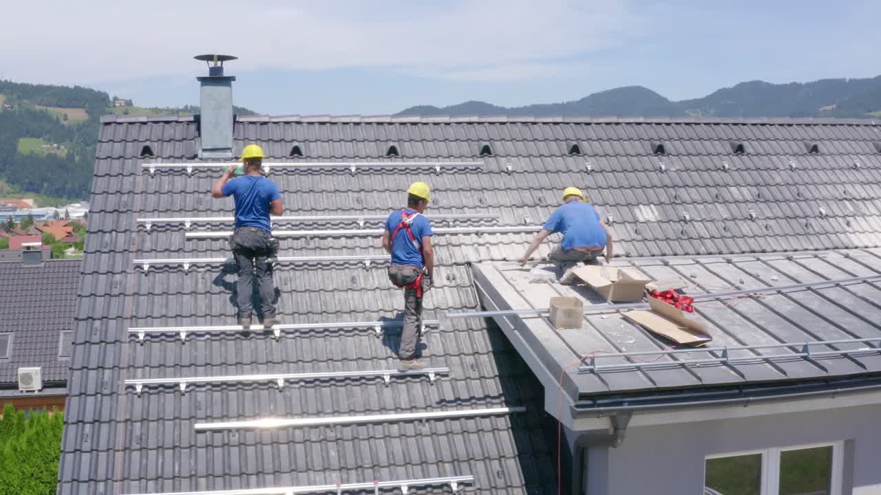 Aerial View Of Three Solar Panel Installers On Roof Of Building. Follow Shot