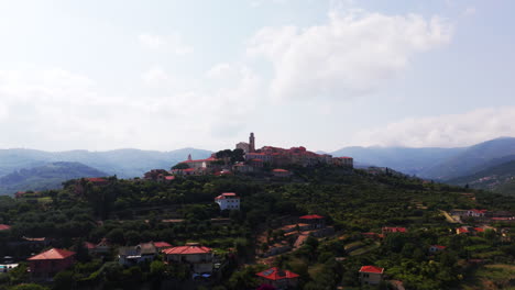 drone approaches a hilltop church with a slight circular move, revealing surrounding village, green vegetation, and mountain landscape in liguria