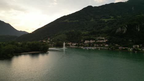 wonderful-nature-with-incredible-natural-habitat-for-people-small-city-town-with-houses-on-a-hill-covered-in-between-trees-boats-in-a-dock-stand-by-great-fountain-releasing-streams-of-water-high-up