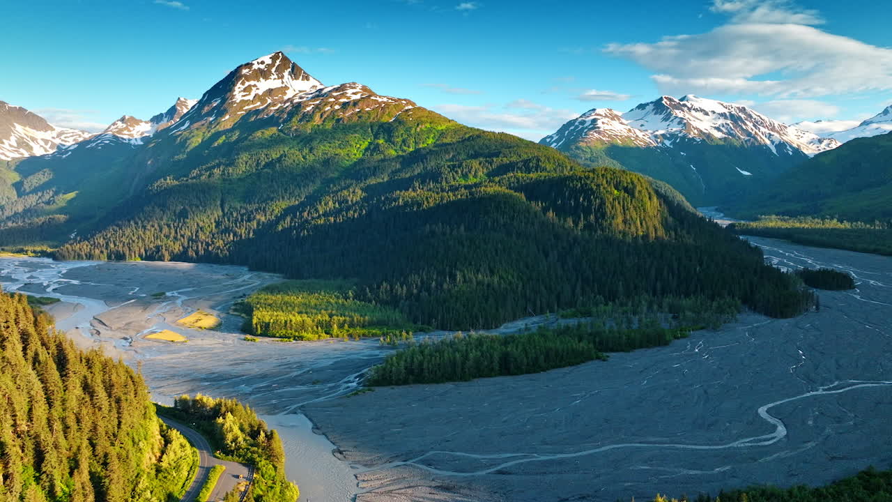 Delta of dried up river flowing among the stunning verdant mountains. Sun lights the tops of the snow-capped rocks. Alaska, USA