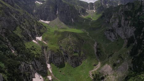 Lush-green-valley-amidst-towering-Bucegi-Mountains-under-a-cloudy-sky,-aerial-view