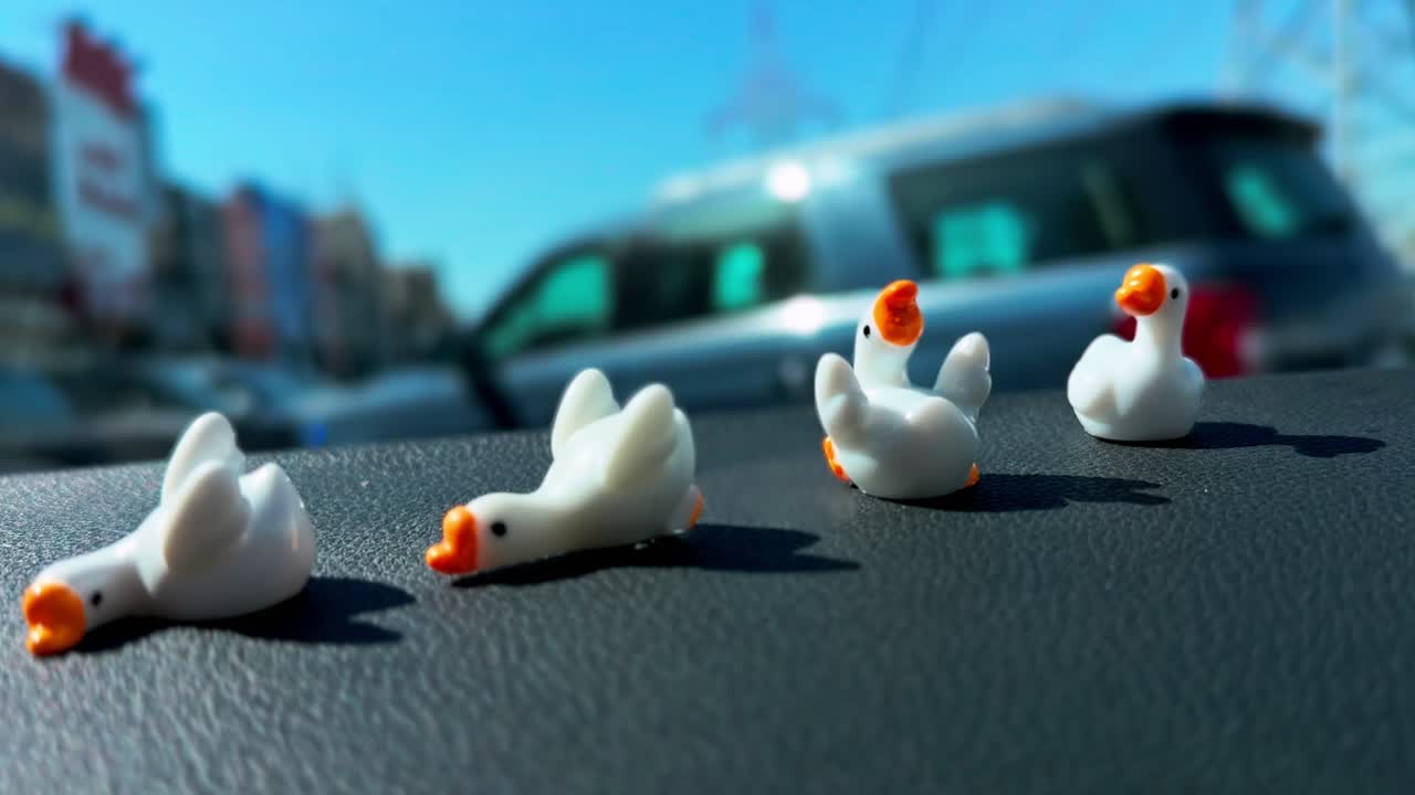 Primo piano con profondità di campo ridotta che mostra piccole statuette di anatre bianche disposte sul cruscotto di un'auto, con strada urbana sfocata e veicolo parcheggiato sotto la luminosa luce solare diurna.