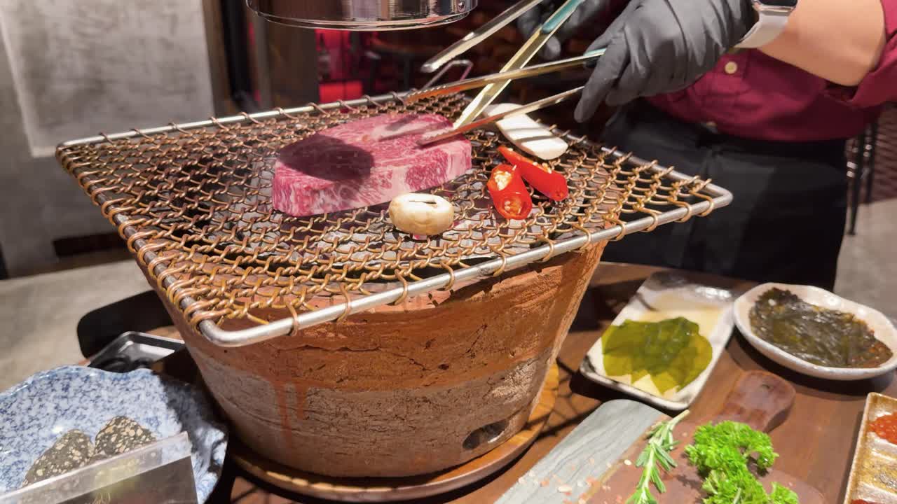 Chef grills thick wagyu steak over charcoal, using tongs and smoke, in restaurant setting