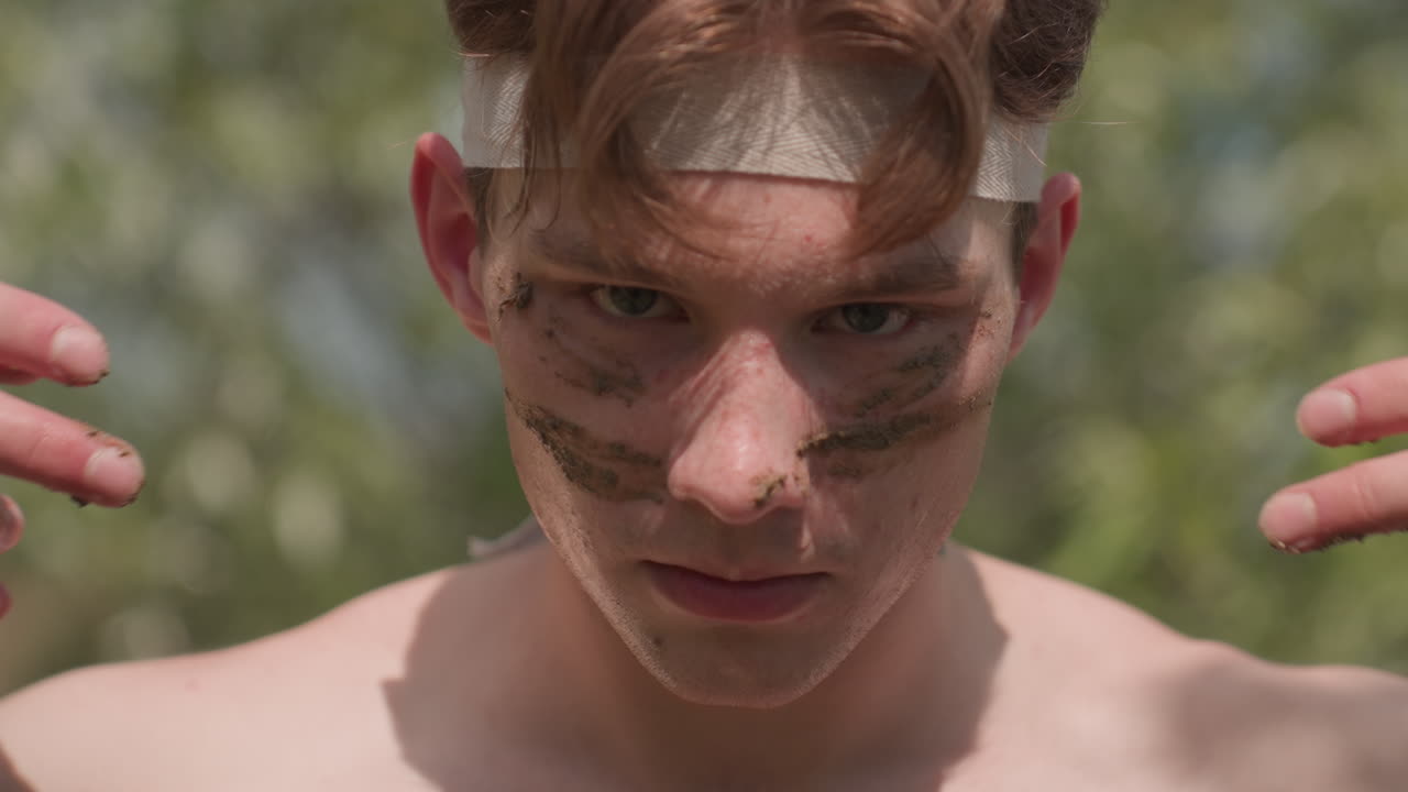 Close-up of shirtless soldier wearing head band, holding wet sand between fingers while preparing to apply it to face as part of intense outdoor ritual surrounded by nature and sunlight