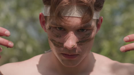 close-up of shirtless soldier wearing head band, holding wet sand between fingers while preparing to apply it to face as part of intense outdoor ritual surrounded by nature and sunlight