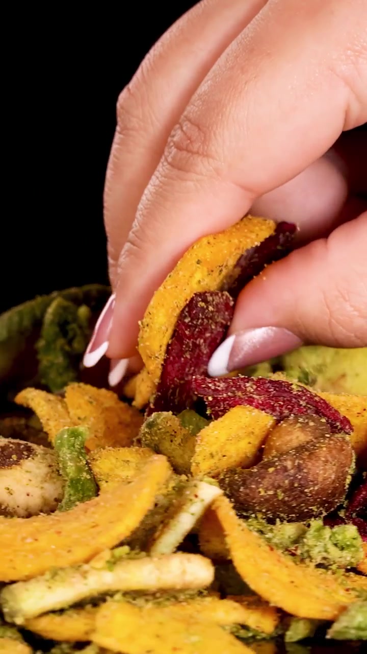 Una mano si allunga e prende patatine croccanti assortite di verdure essiccate, inclusi fagioli, barbabietola, patate e funghi, sotto una luminosa illuminazione da studio con uno sfondo nero