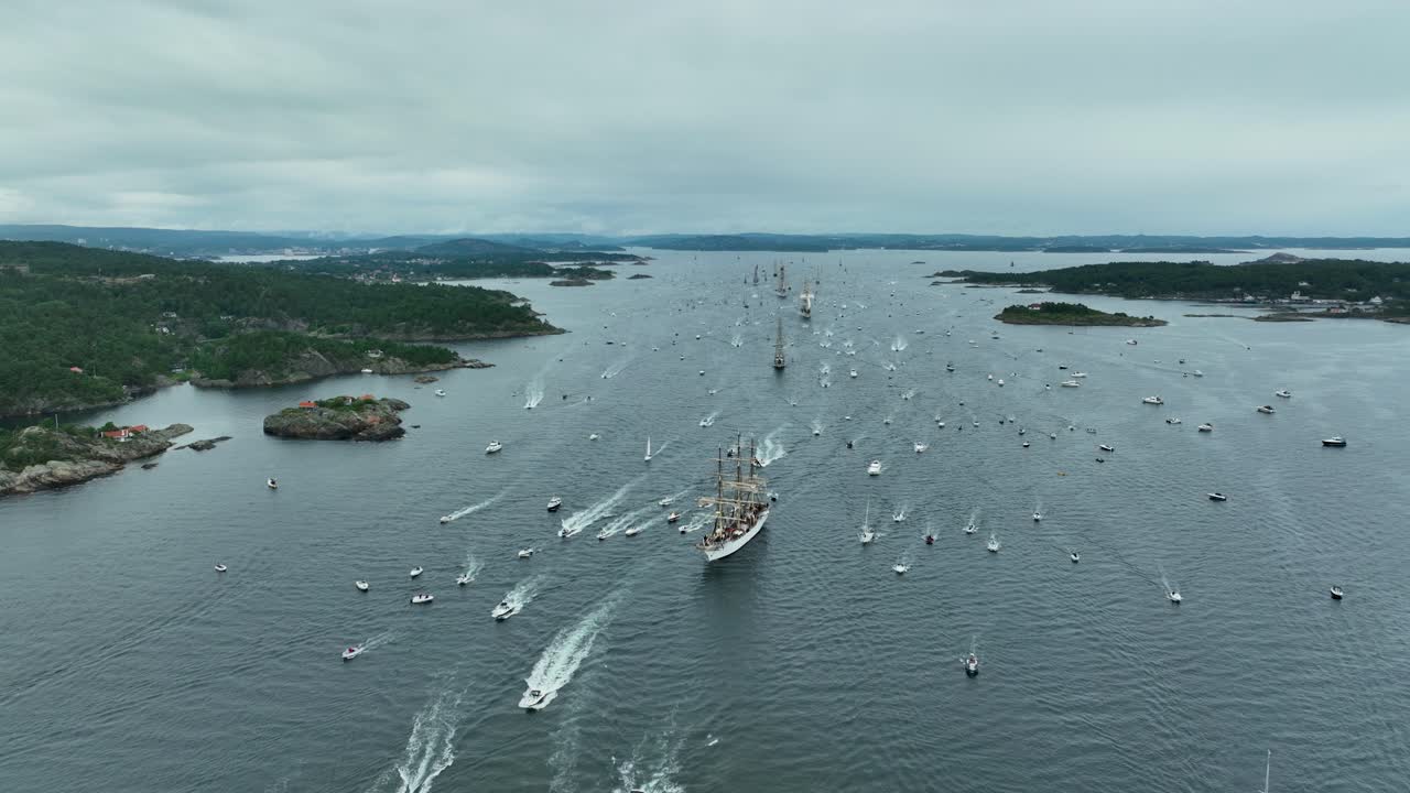 An array of boats and ships navigates through tranquil waters, creating ripples as they venture among scenic islands under overcast skies during a maritime festival