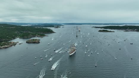 an array of boats and ships navigates through tranquil waters, creating ripples as they venture among scenic islands under overcast skies during a maritime festival
