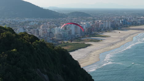 cinematic establishing wide angle aerial shot following a red kite paraglider flying towards caioba beach, brazil