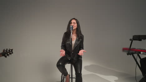 una hermosa vocalista con una chaqueta de cuero negra, sonriendo y cantando con los ojos abiertos y las manos moviéndose expresivamente, de pie en un micrófono vintage en un taburete alto, actuando contra un fondo gris
