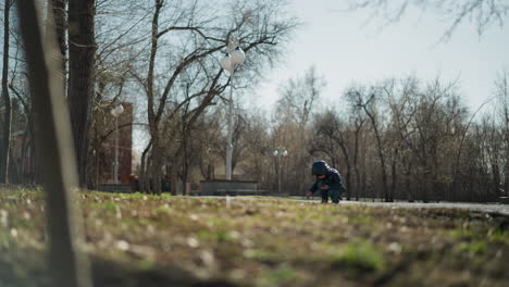 a young boy, dressed warmly in a black leather jacket, head warmer, and jeans, is squatting in an open area while focused on digging into the ground with a stick